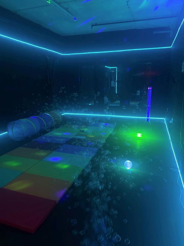 Sensory room in calm teal light, with soft mats, a play tunnel and bubbles floating past a glowing green light