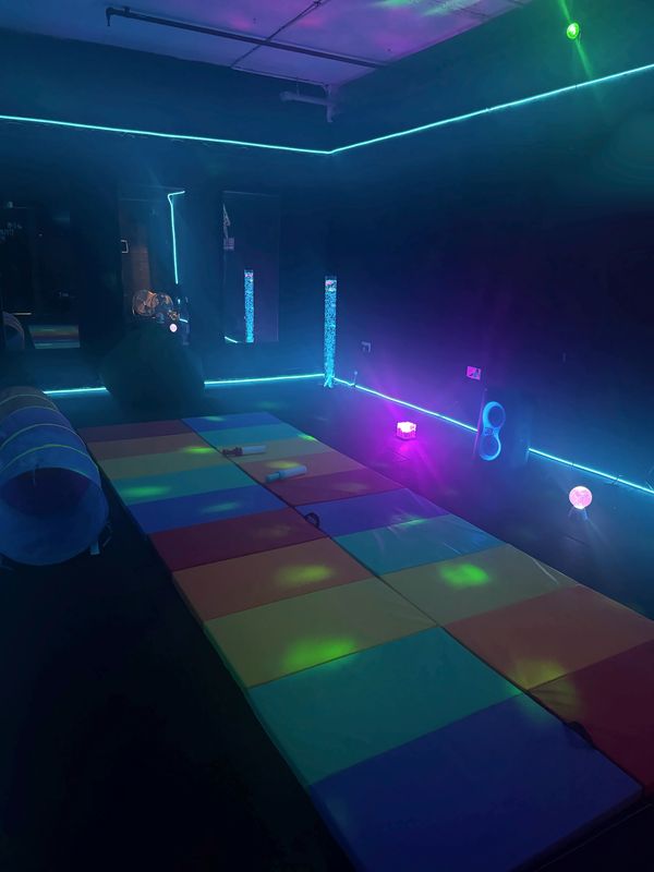 Wide view of the sensory room in blue light, with a long rainbow path of soft mats and the play tunnel